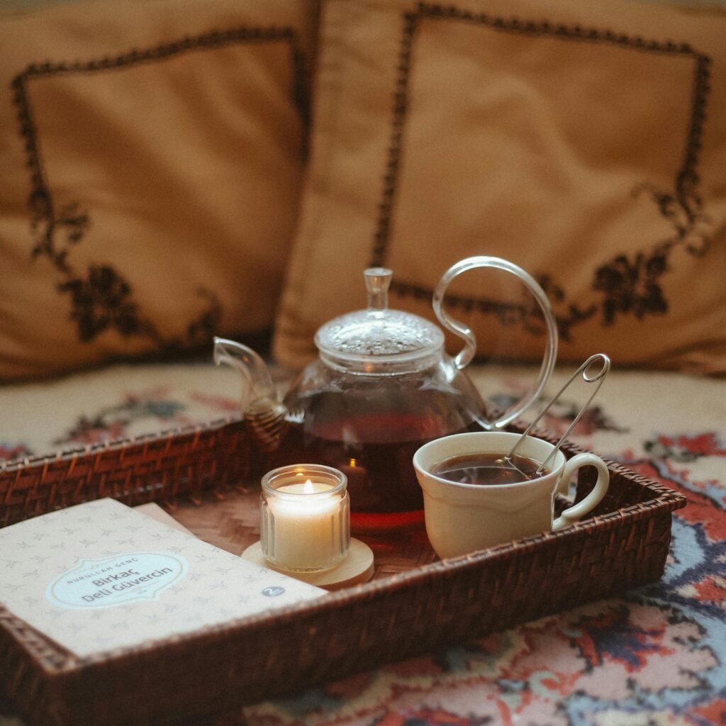 A warm, relaxing setup with a teapot, tea cup, candle, and book on a wicker tray.