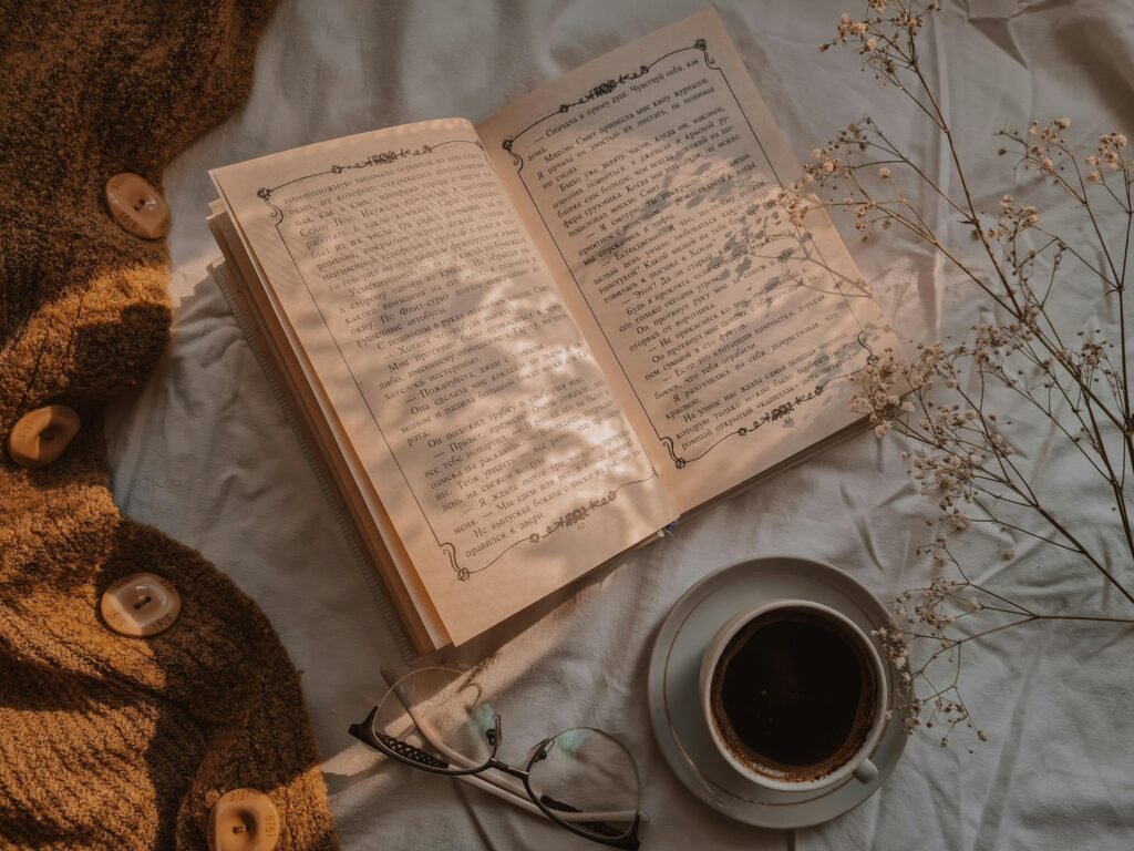 Warm flat lay image featuring an open book, coffee, and dried flowers on a cozy fabric background.